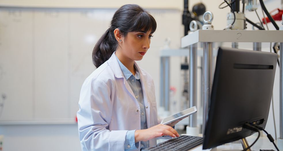 Image shows a woman in a white lab coat using a computer in a laboratory setting.