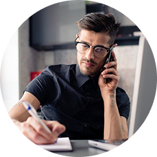 A man is taking notes while speaking on a phone at his desk.
